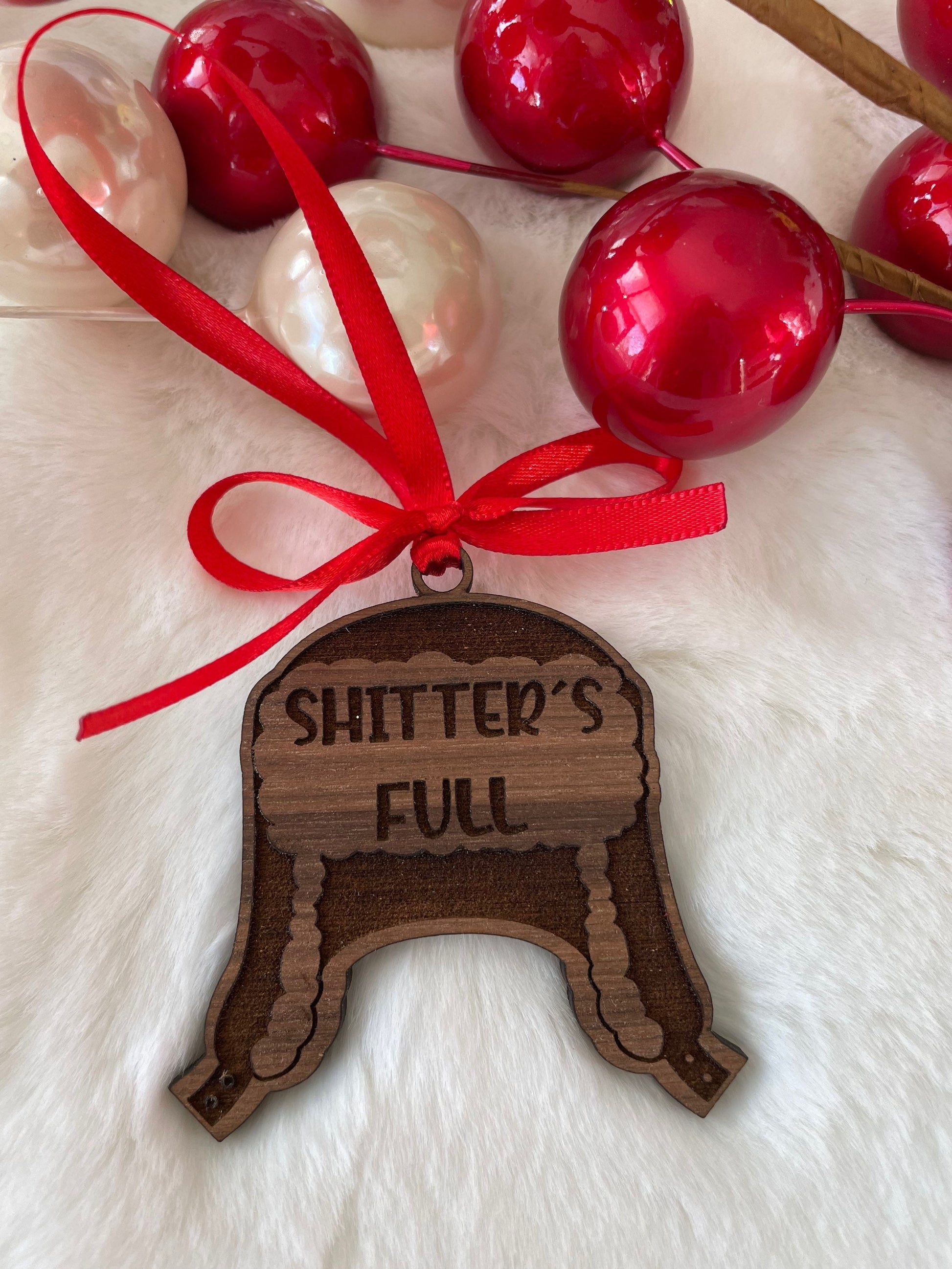Wooden ornament with 'SHITTER'S FULL' text on a white surface with red and white ornaments.